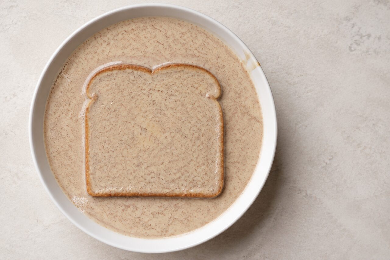A square bread slice lying flat and fully submerged in pale beige speckled custard in a wide shallow bowl, seen from directly overhead