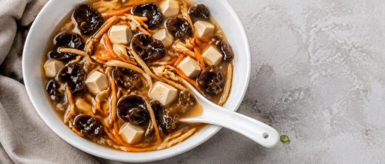 An overhead view of a wide white bowl packed with dark wood ear curls, carrot matchsticks, torn tofu and pale strands in a translucent brown broth