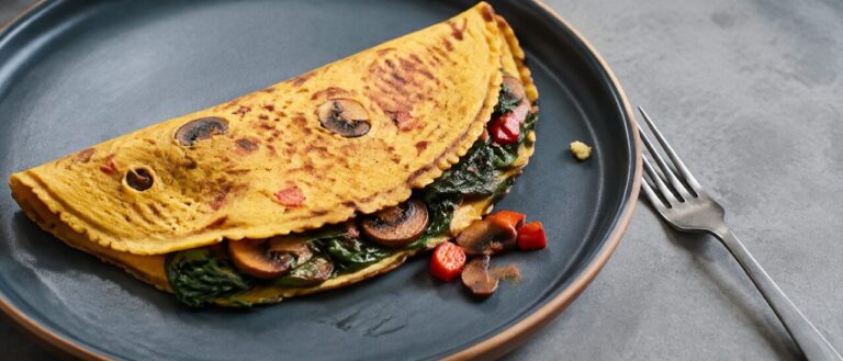 A folded chickpea flour omelette on a matte stoneware plate with wilted spinach and sliced mushrooms showing at the open edge